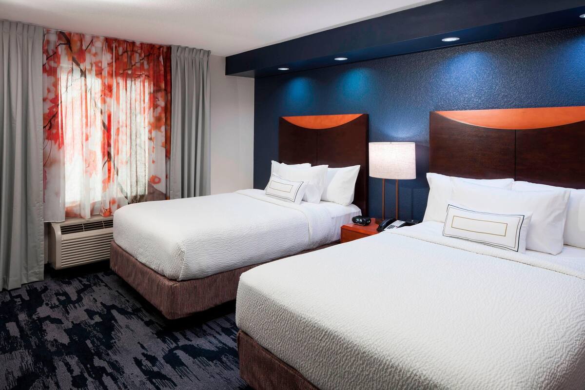 A hotel room with two beds, a large window with sheer curtains, a wall-mounted air conditioner, and a nightstand with a lamp.