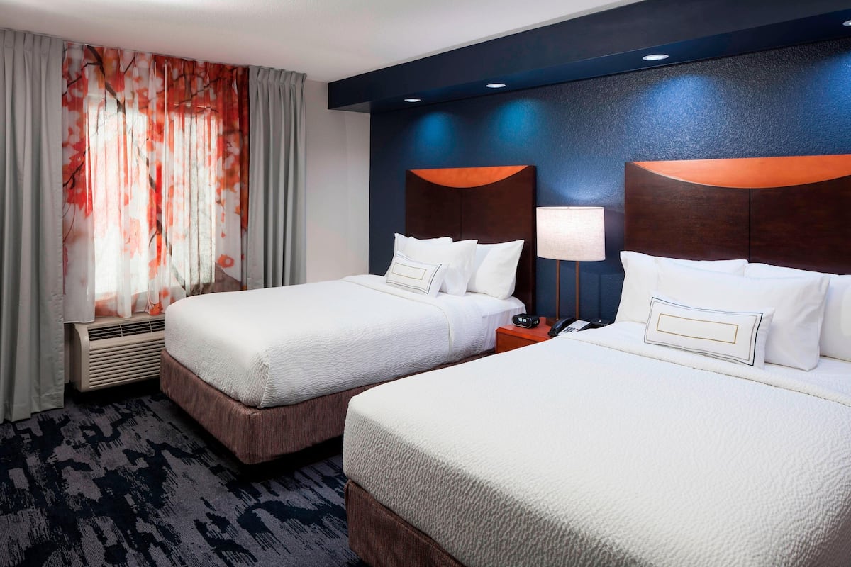 A hotel room with two beds, a large window with sheer curtains, a wall-mounted air conditioner, and a nightstand with a lamp.