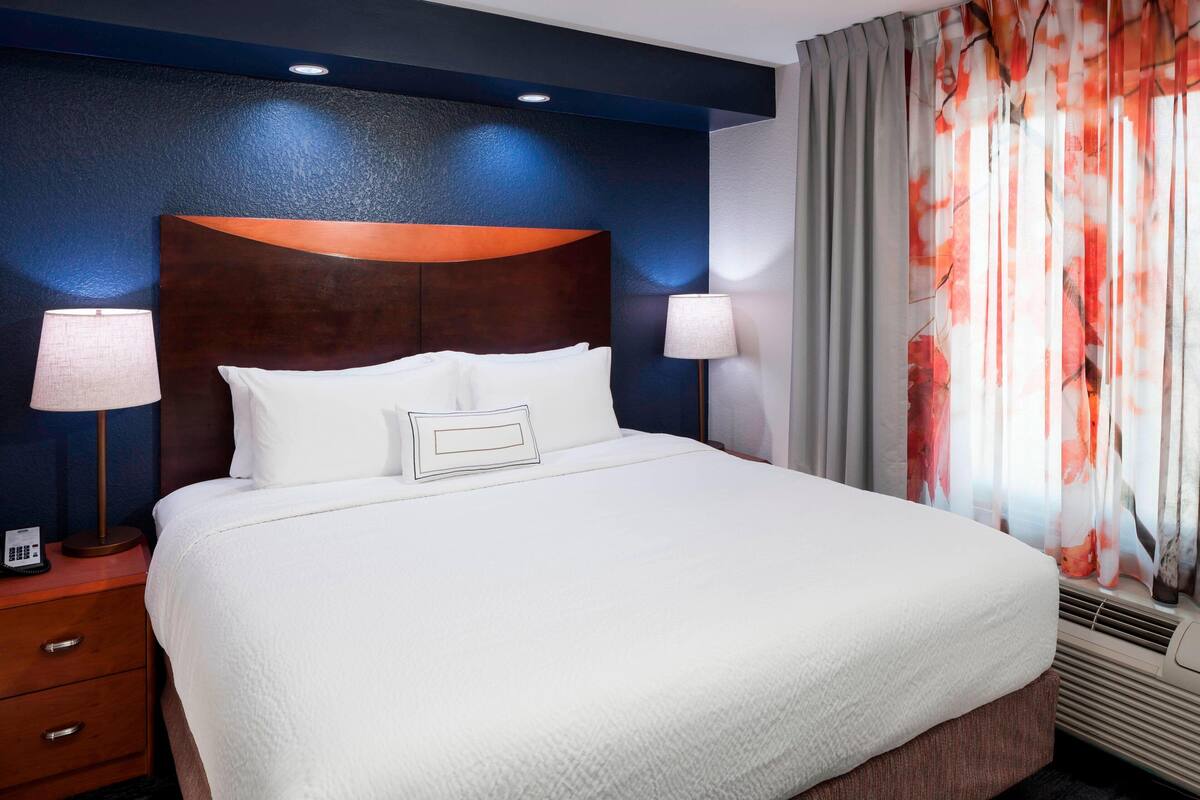 A hotel room with a large bed, two bedside lamps, a telephone on the nightstand, and a window with curtains.
