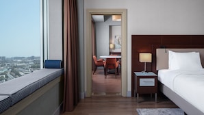Ein Hotelzimmer mit großem Fenster, einem Bett, einem Nachttisch, einem Schreibtisch mit Stühlen und Blick auf die Stadt.