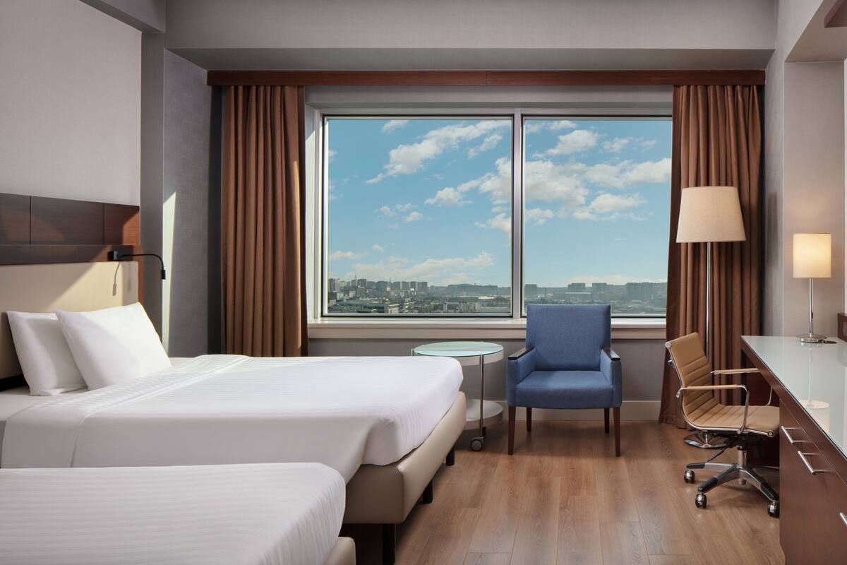 A hotel room with two beds, a desk, a chair, a lamp, and a view of the city through large windows.