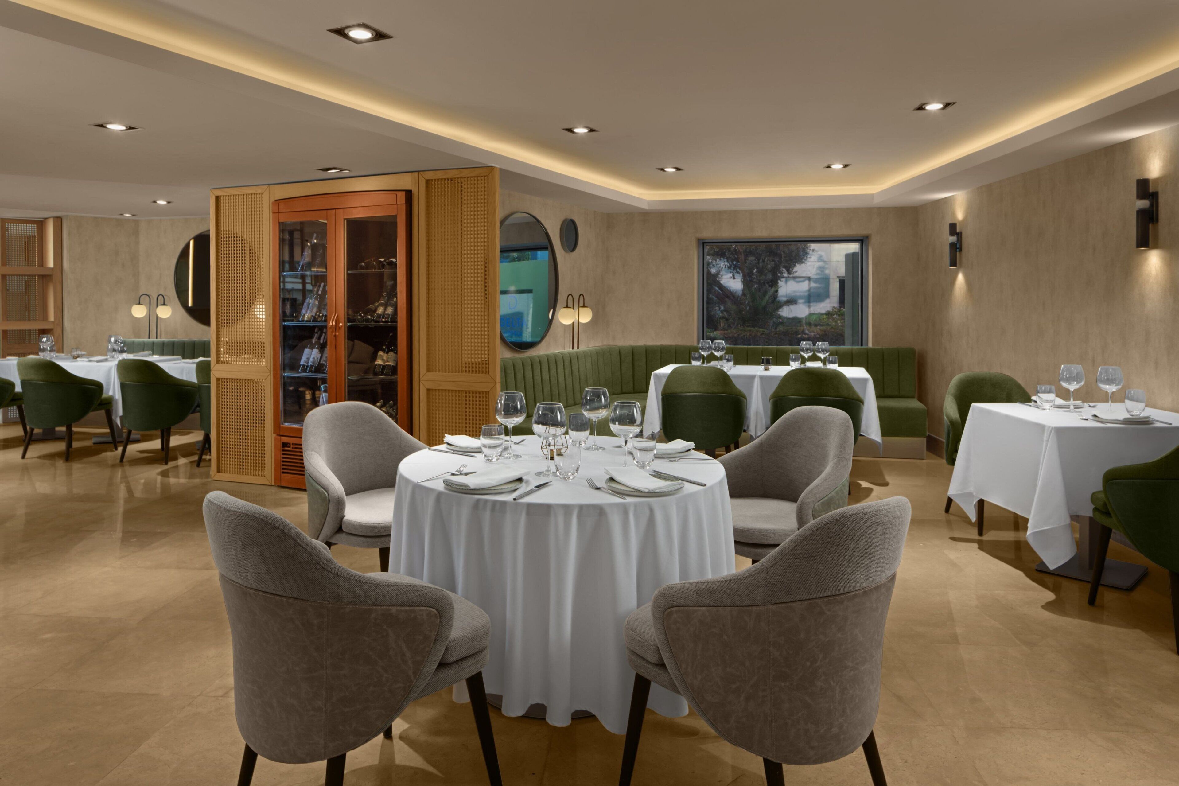 A restaurant with round tables, green upholstered chairs, and a large window with a view.