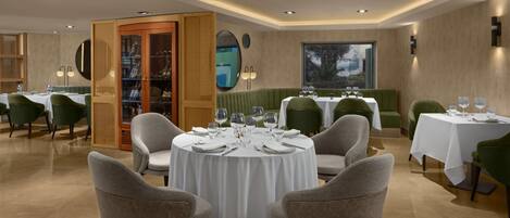 A restaurant with round tables, green upholstered chairs, and a large window with a view.