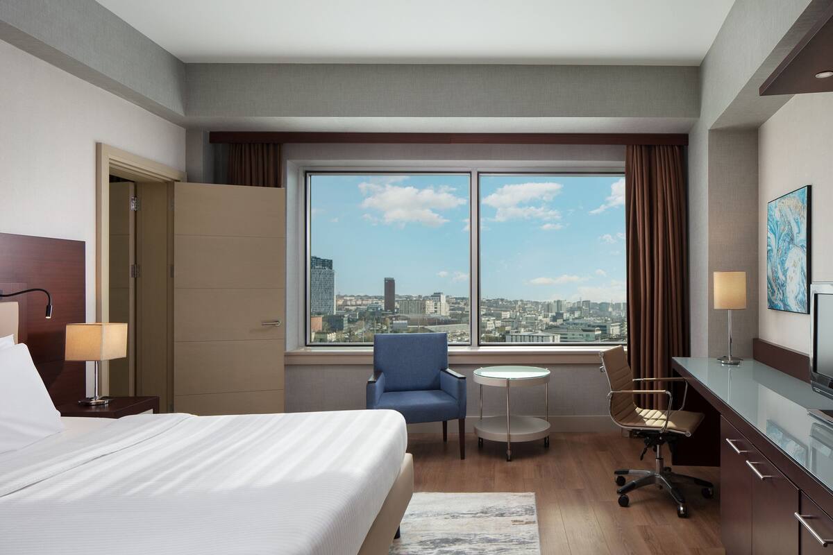 A hotel room with a bed, a desk, a chair, a small table, and a view of the city through large windows.