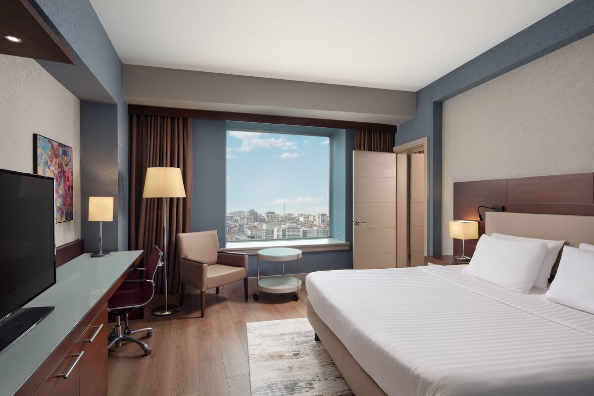 A hotel room with a bed, desk, chair, TV, and a window with a city view.