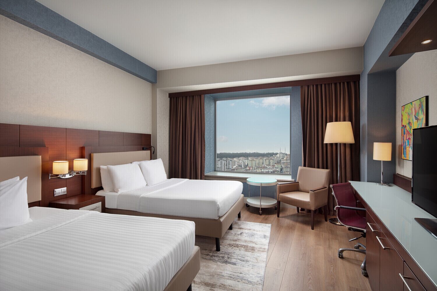 A hotel room with two beds, a desk, a chair, a TV, and a view of a cityscape.