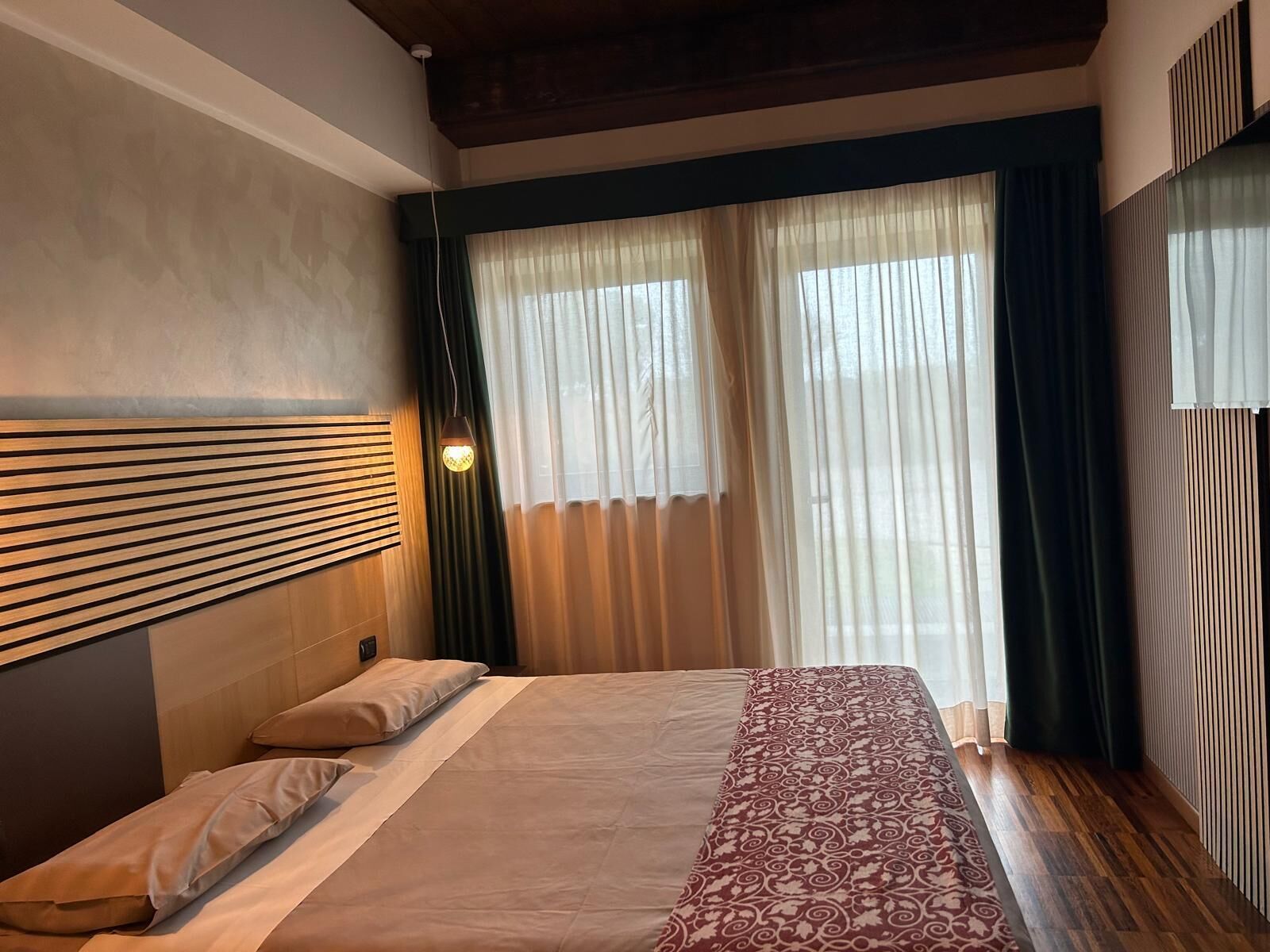 A hotel room with a bed, wooden headboard, and a large window with sheer curtains.