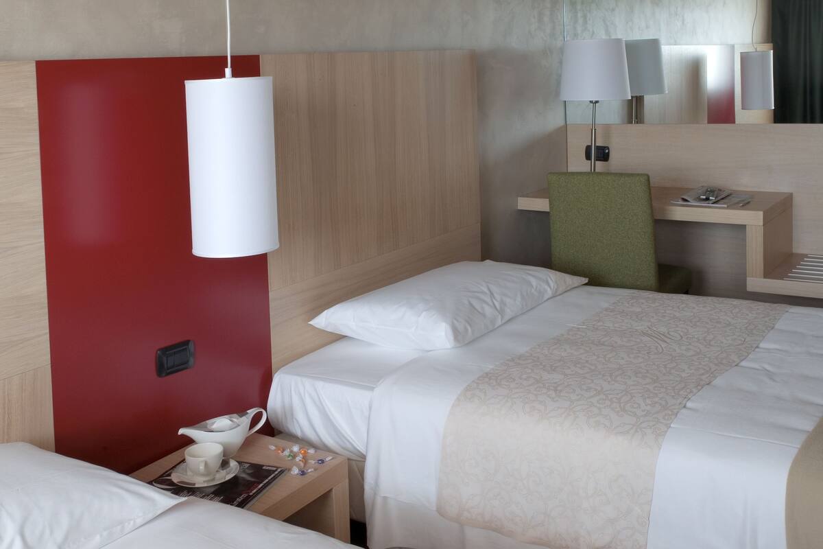 A hotel room with two beds, a desk, a chair, and a bedside table with a lamp.