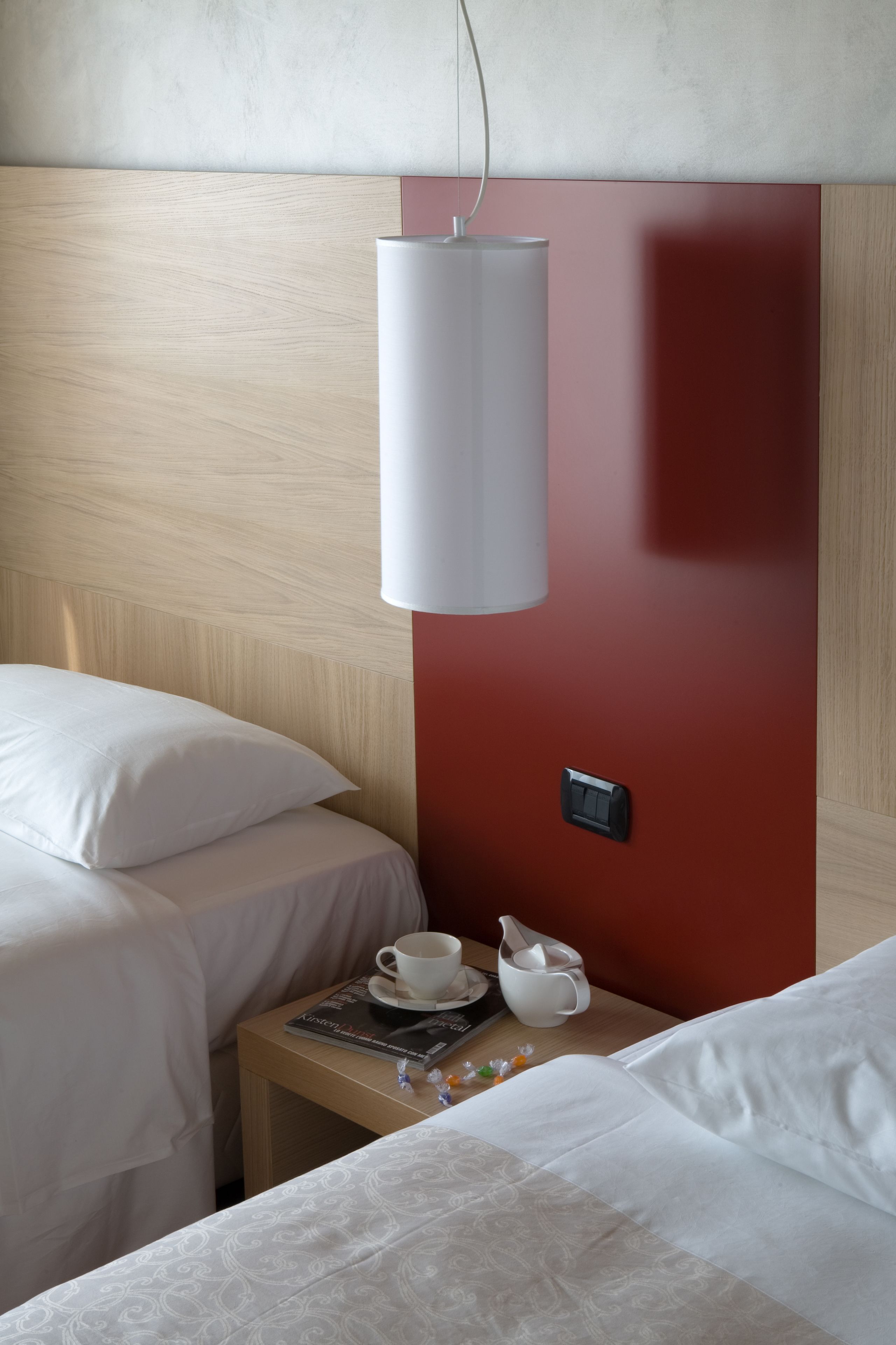 A hotel room with a wooden headboard, a red accent wall, a bedside table with a cup, a book, and a remote control.