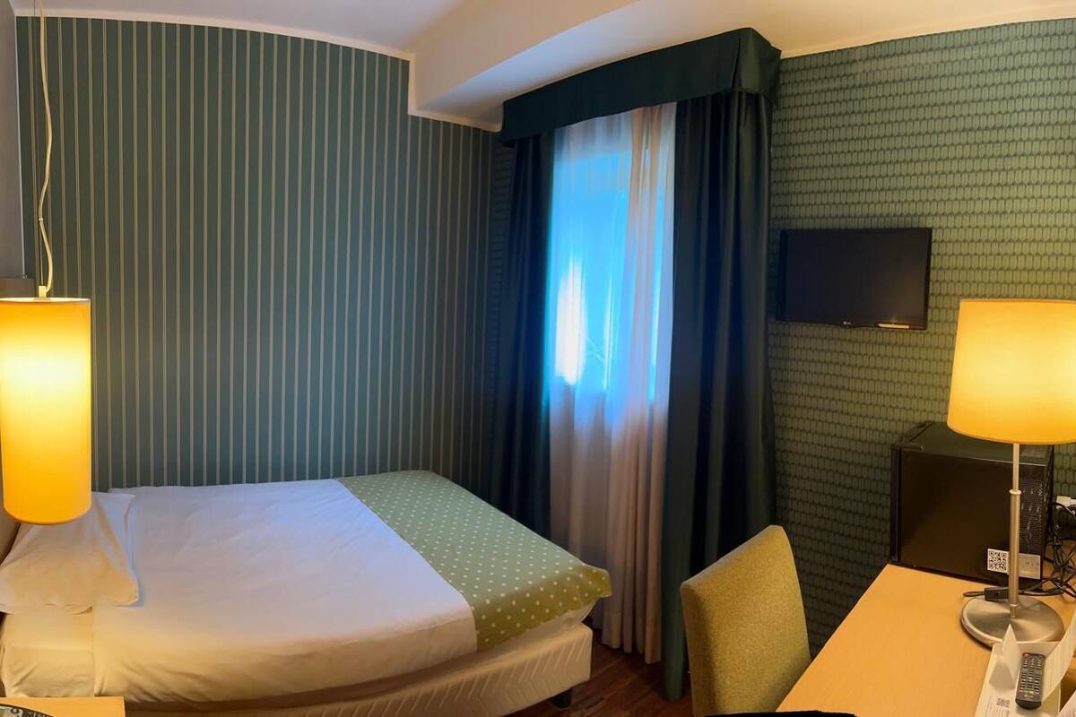 A hotel room with a bed, two bedside lamps, a TV, and a desk with a lamp.