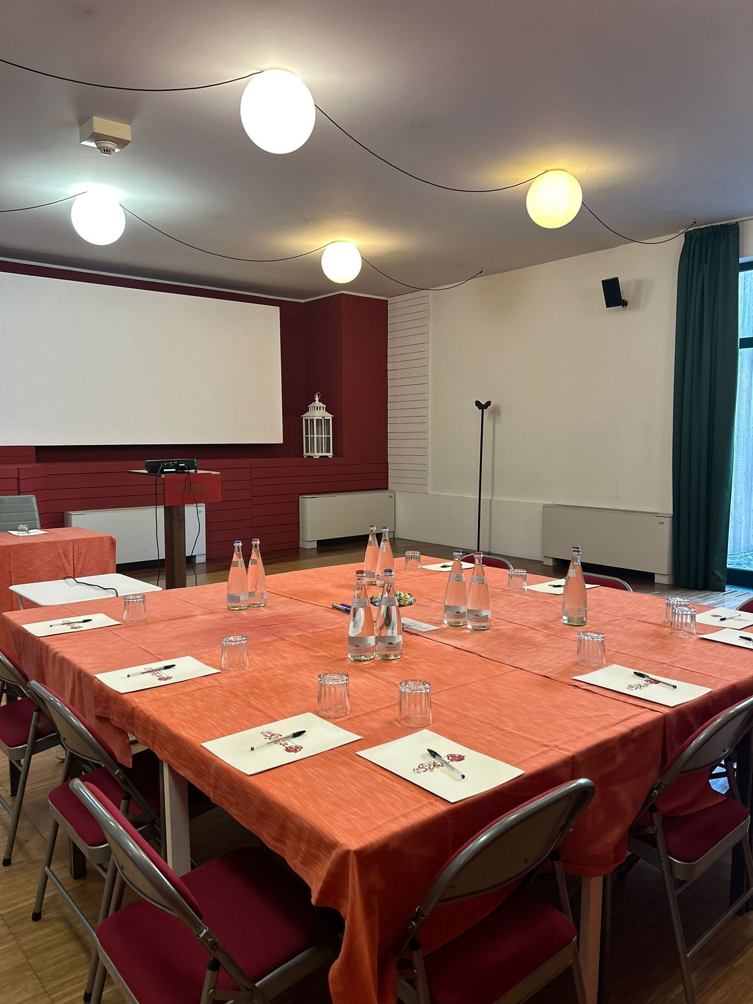 A conference room with a long table, red chairs, water bottles, and a projector screen.