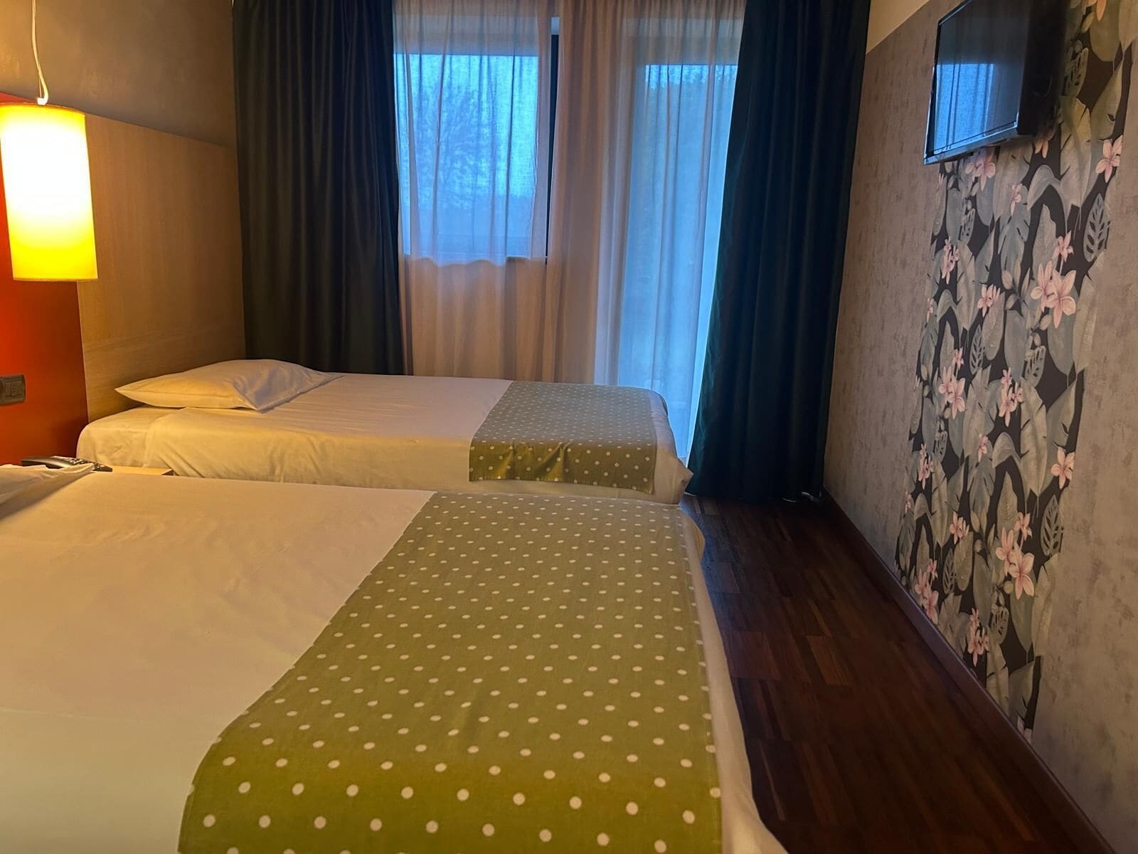 A hotel room with two beds, a TV mounted on the wall, and a window with curtains.