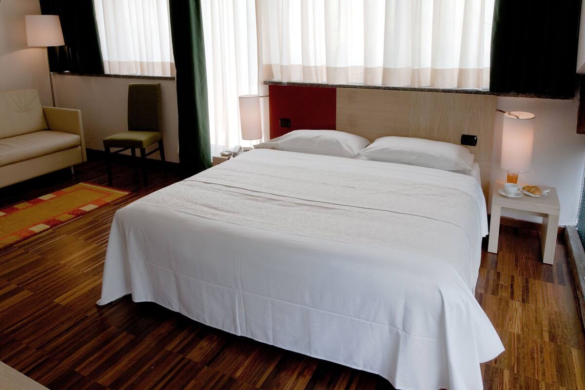 A hotel room with a large bed, a chair, a small table with a lamp, and a bedside table with a phone and a glass of water.