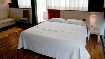 Hotelkamer met een groot bed, een stoel, een klein tafeltje met een lamp en een nachtkastje met een telefoon en een glas water.