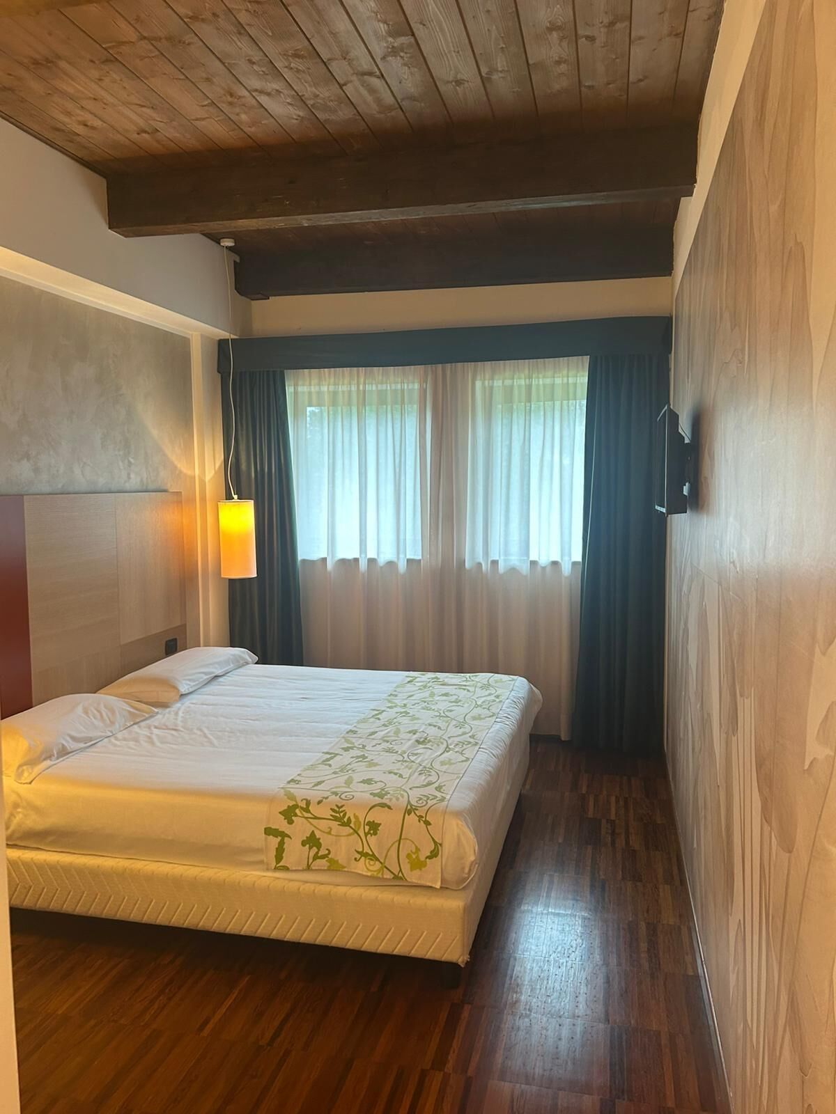 A hotel room with a wooden ceiling, a bed with a white mattress and green floral bedspread, a bedside lamp, and a window with curtains.