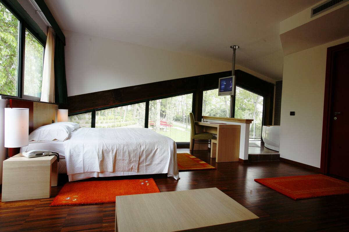 A modern bedroom with a large bed, desk, and chair, a small table, and a view of greenery outside.