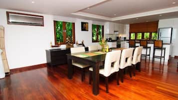 A spacious dining area with a wooden table and chairs, a kitchen in the background, and large windows with a view of greenery.