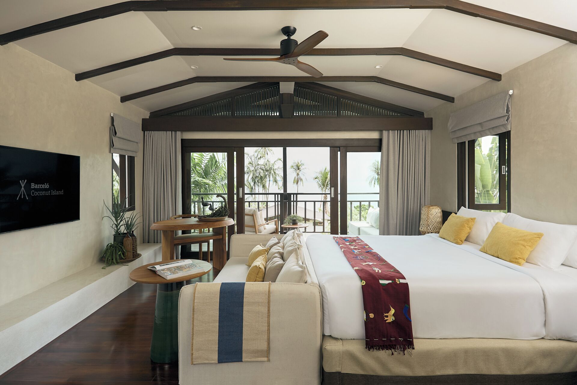 A spacious bedroom with a large bed, a ceiling fan, and a balcony with a view of palm trees.