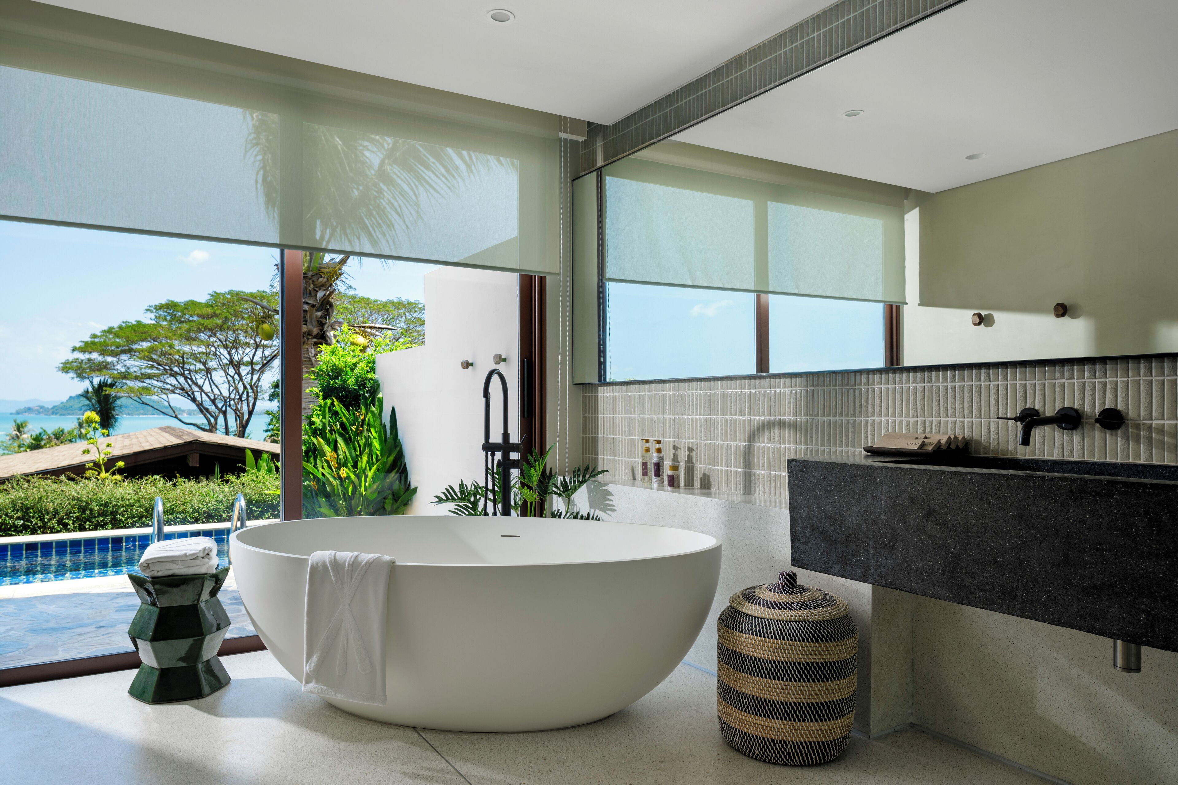 A modern bathroom with a freestanding bathtub, a large mirror, and a view of a pool and tropical landscape.
