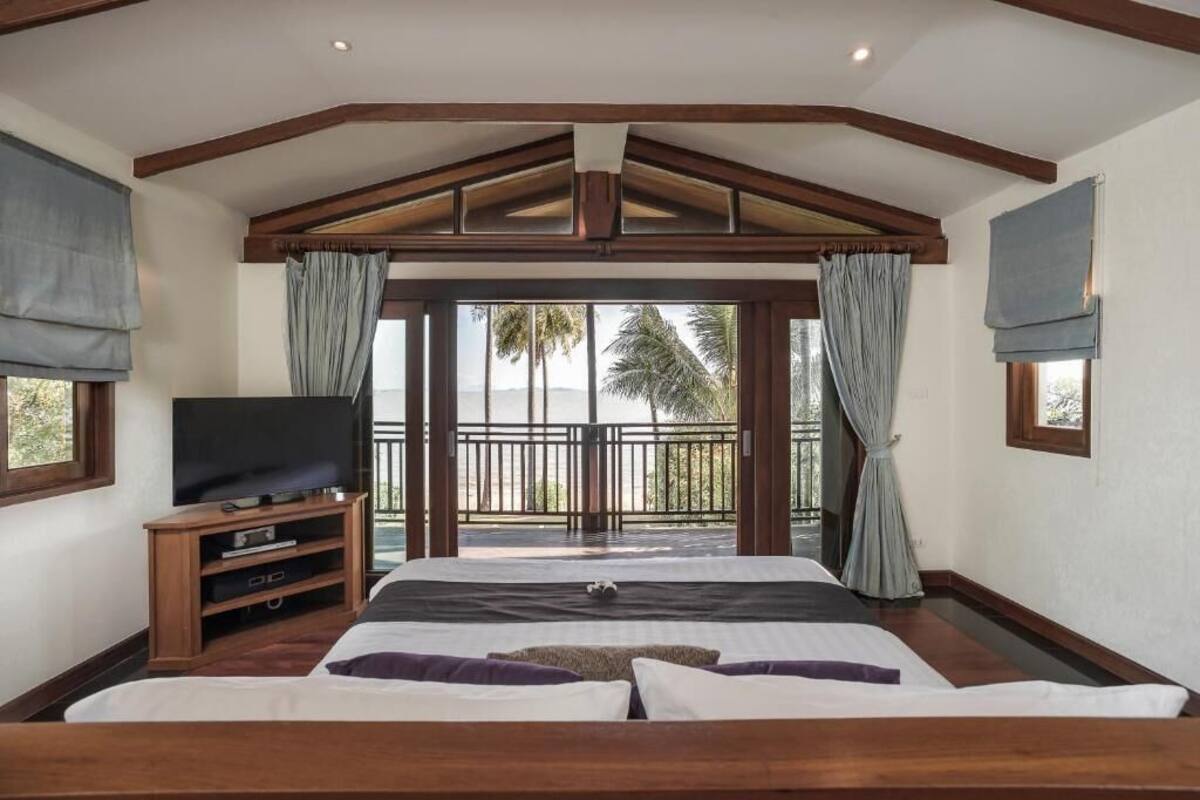 A bedroom with a large bed, a TV, and a balcony with a view of palm trees.