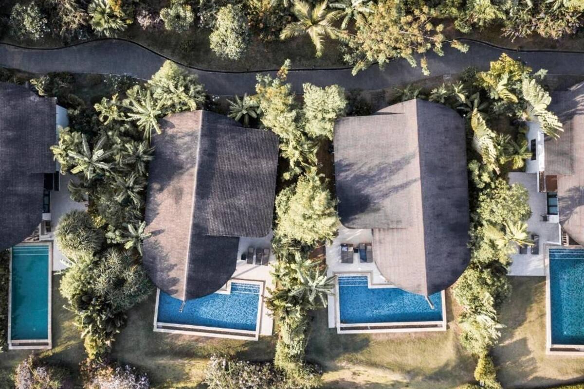 An aerial view of a property with two large, dark-roofed buildings surrounded by lush greenery and two swimming pools.