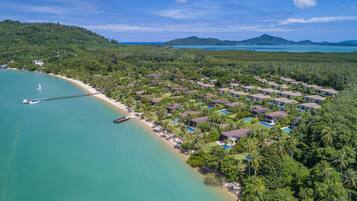 An aerial view of a coastal resort with multiple bungalow-style accommodations, a pier extending into the turquoise sea, and lush greenery surrounding the property.