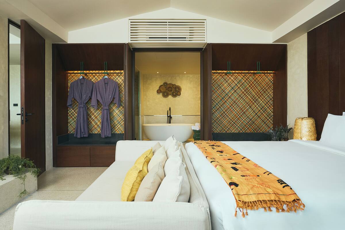A modern bedroom with a large bed, a built-in bench with robes, a vanity with a sink, and patterned walls.