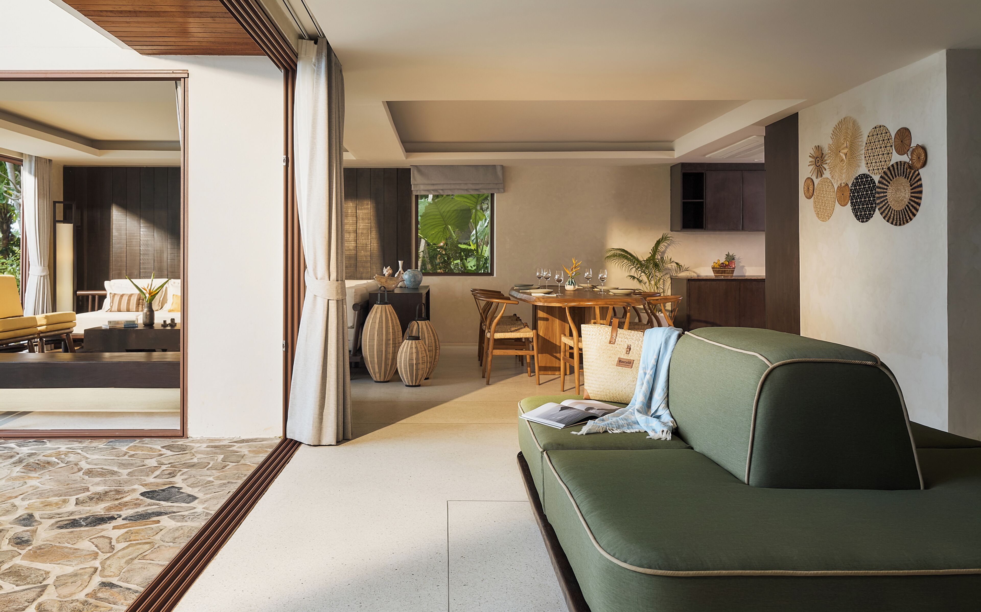 A modern living room with a green sofa, dining table, and chairs. There is a kitchen area with cabinets and a window.