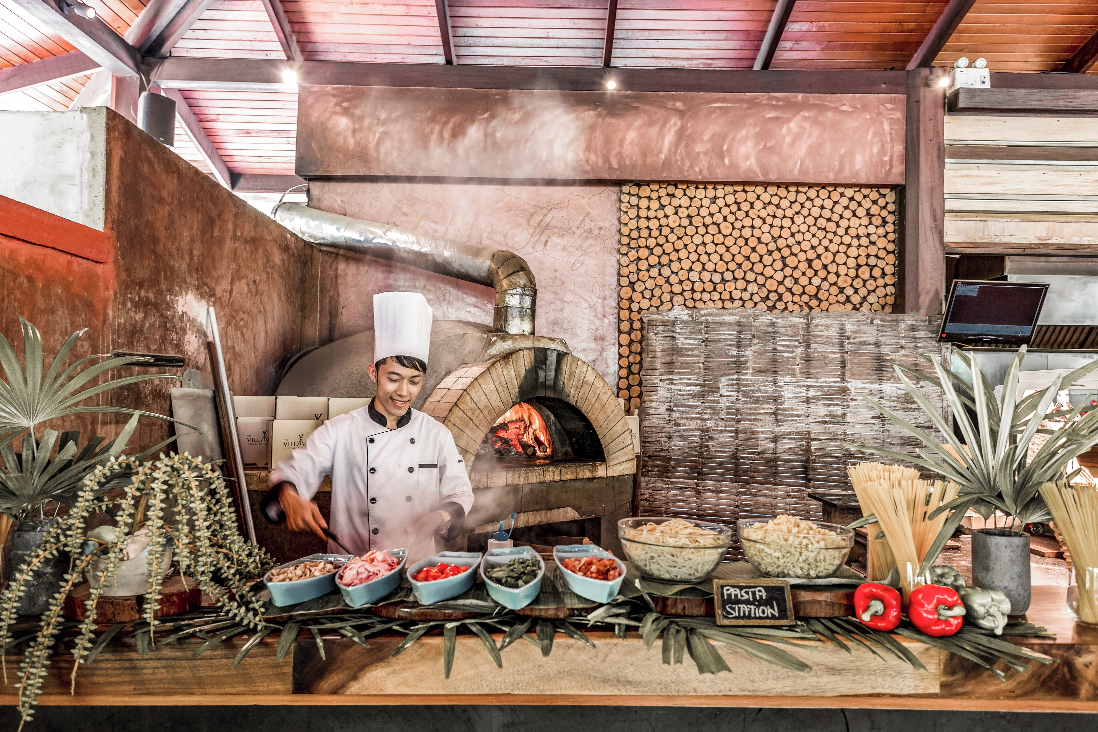 A chef preparing food in front of a wood-fired oven, with various pasta dishes and fresh vegetables displayed on a counter.