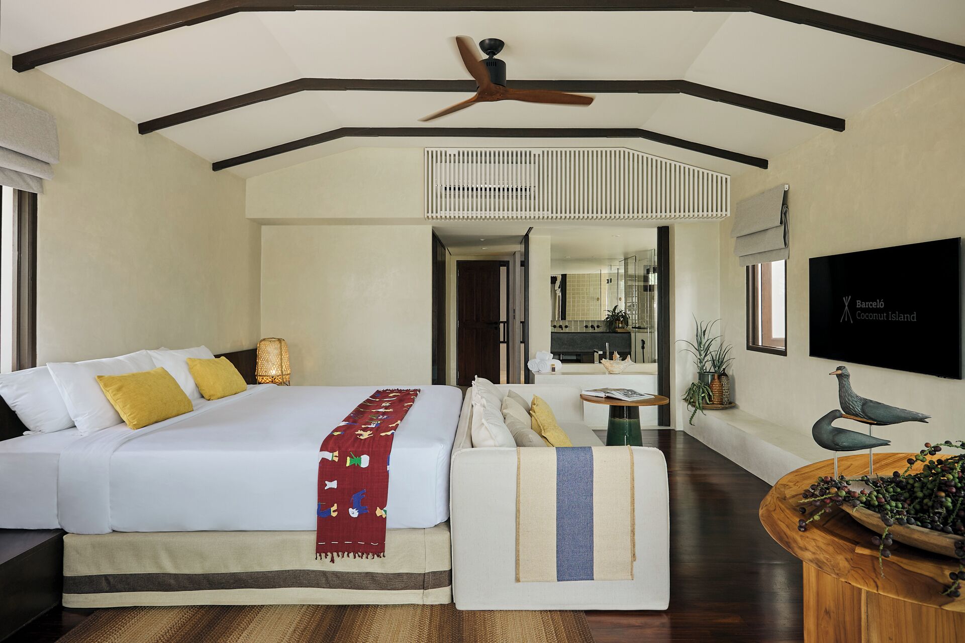 Une chambre d’hôtel avec un grand lit, un canapé, une table d’appoint en bois sur laquelle se trouve un panier de fruits, un ventilateur de plafond et une vue sur la salle de bain et la cuisine.