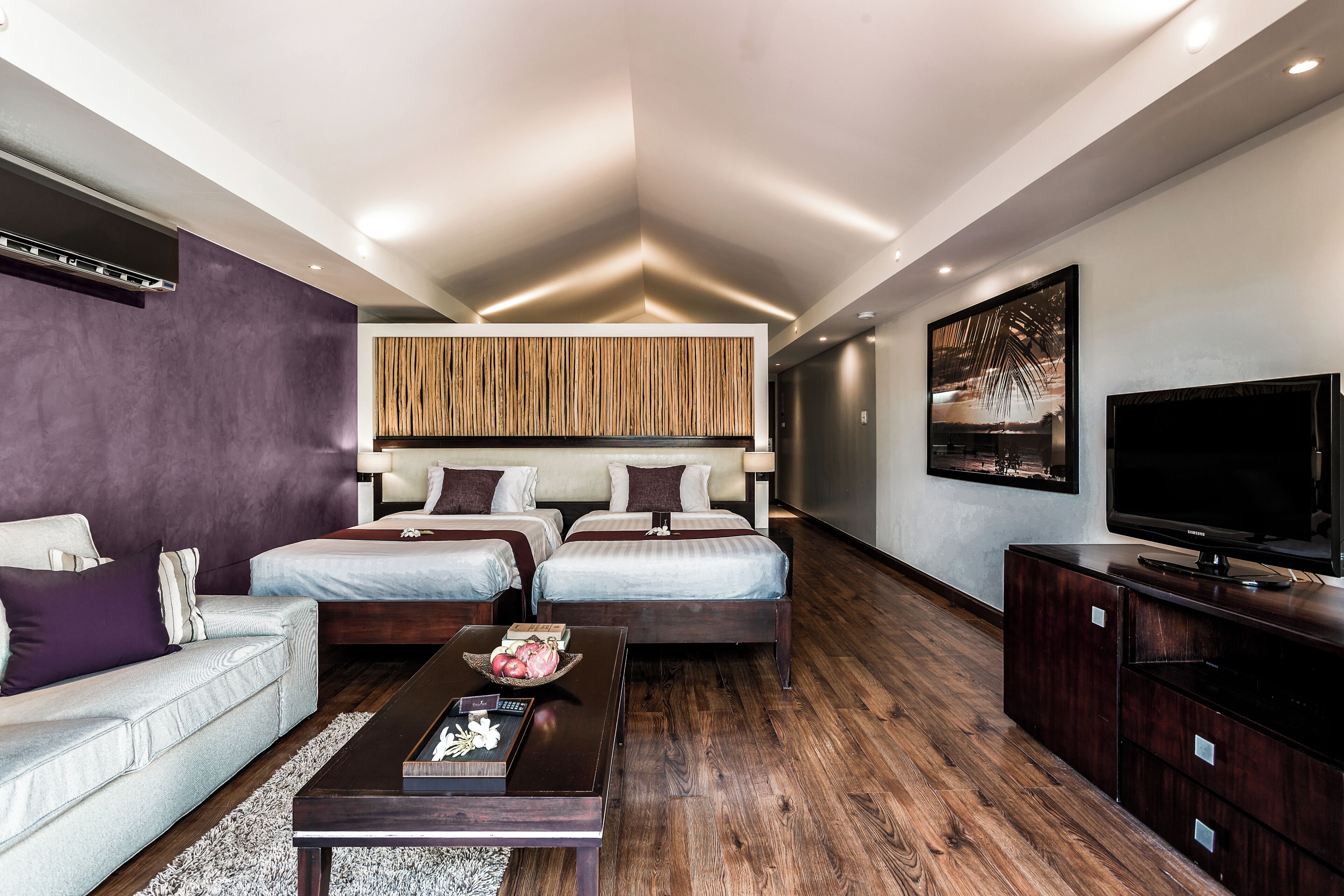 A modern hotel room with a sofa, two beds, a TV, and a wooden floor.