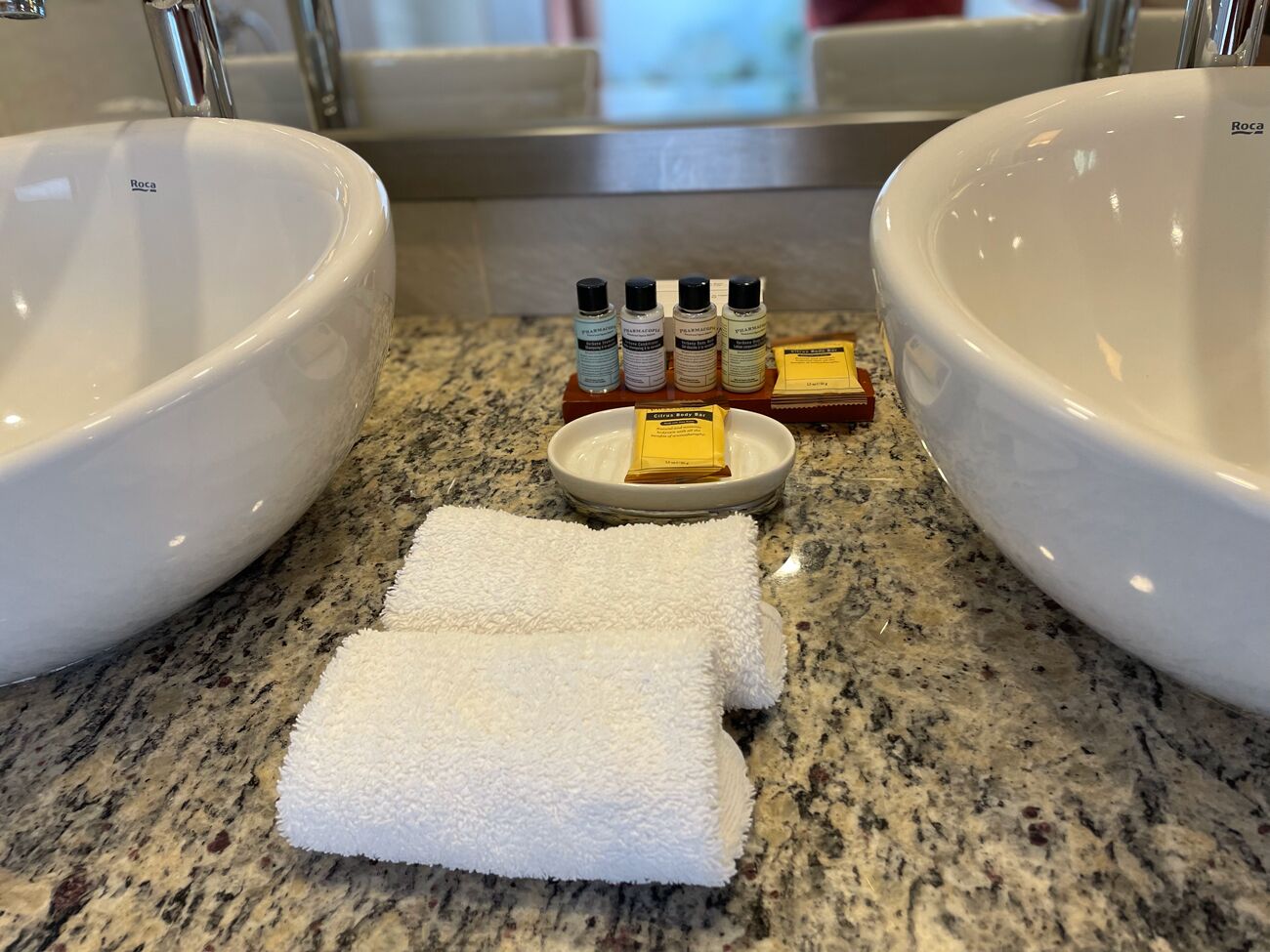 Two white sinks with a granite countertop, a white towel, and toiletries including a soap bar and bottles.