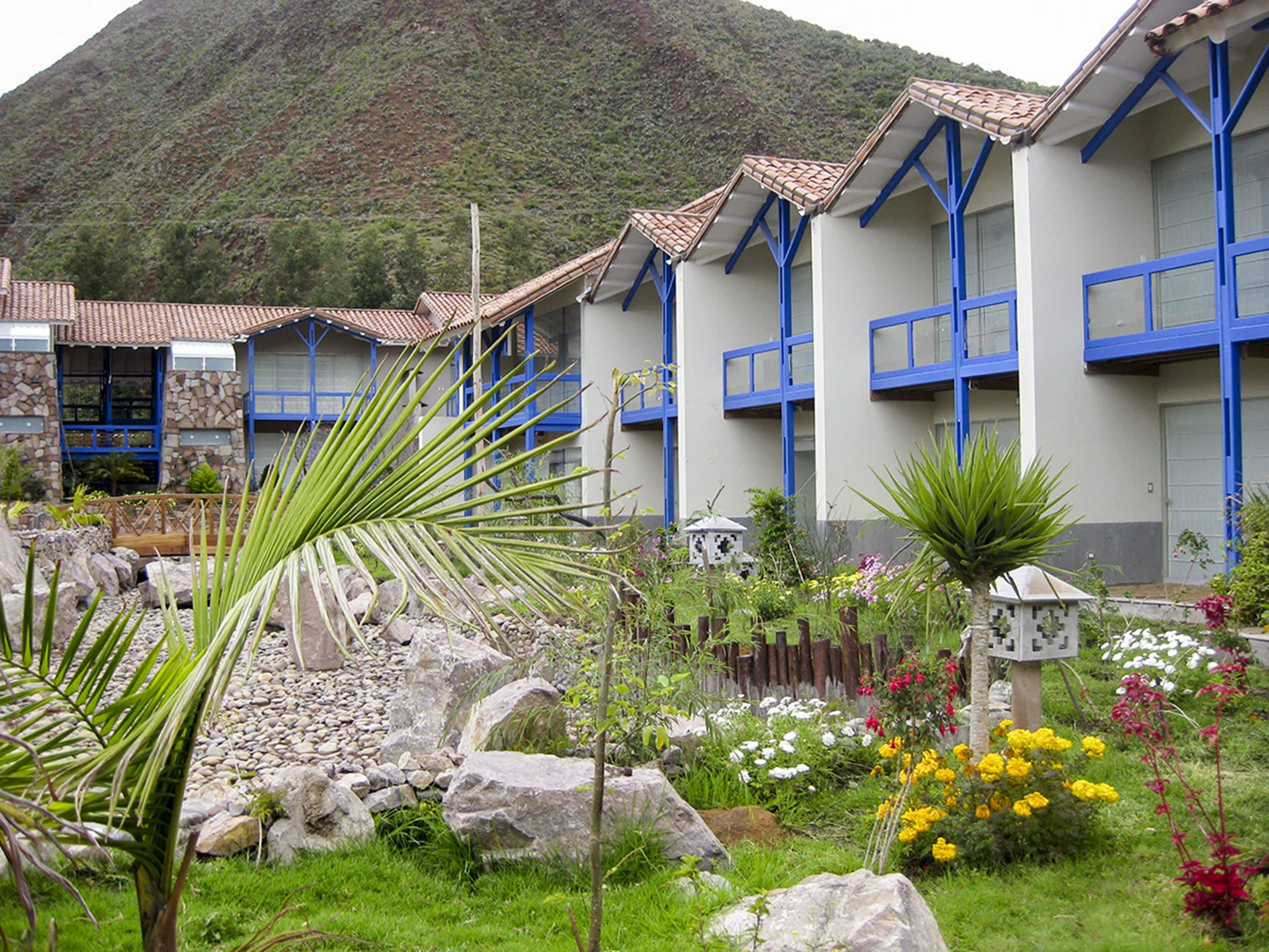 A hotel with multiple blue-framed rooms, a garden with rocks and plants, and a mountainous backdrop.