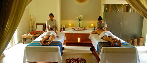 A spa room with two massage tables, a central massage table, and a small table with a vase of flowers.