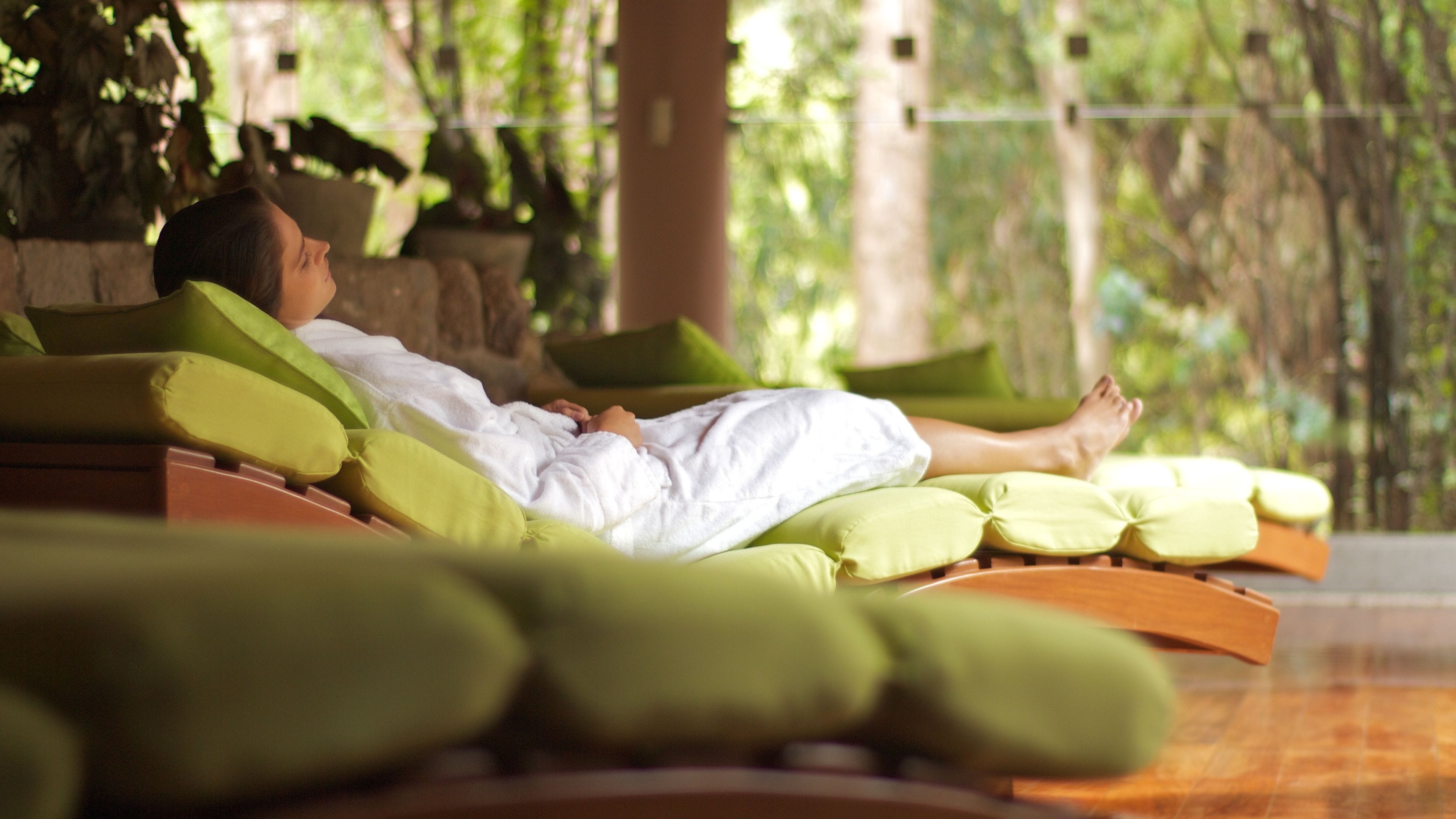 A person lying on a sun lounger with green cushions, in a room with a stone wall and greenery in the background.