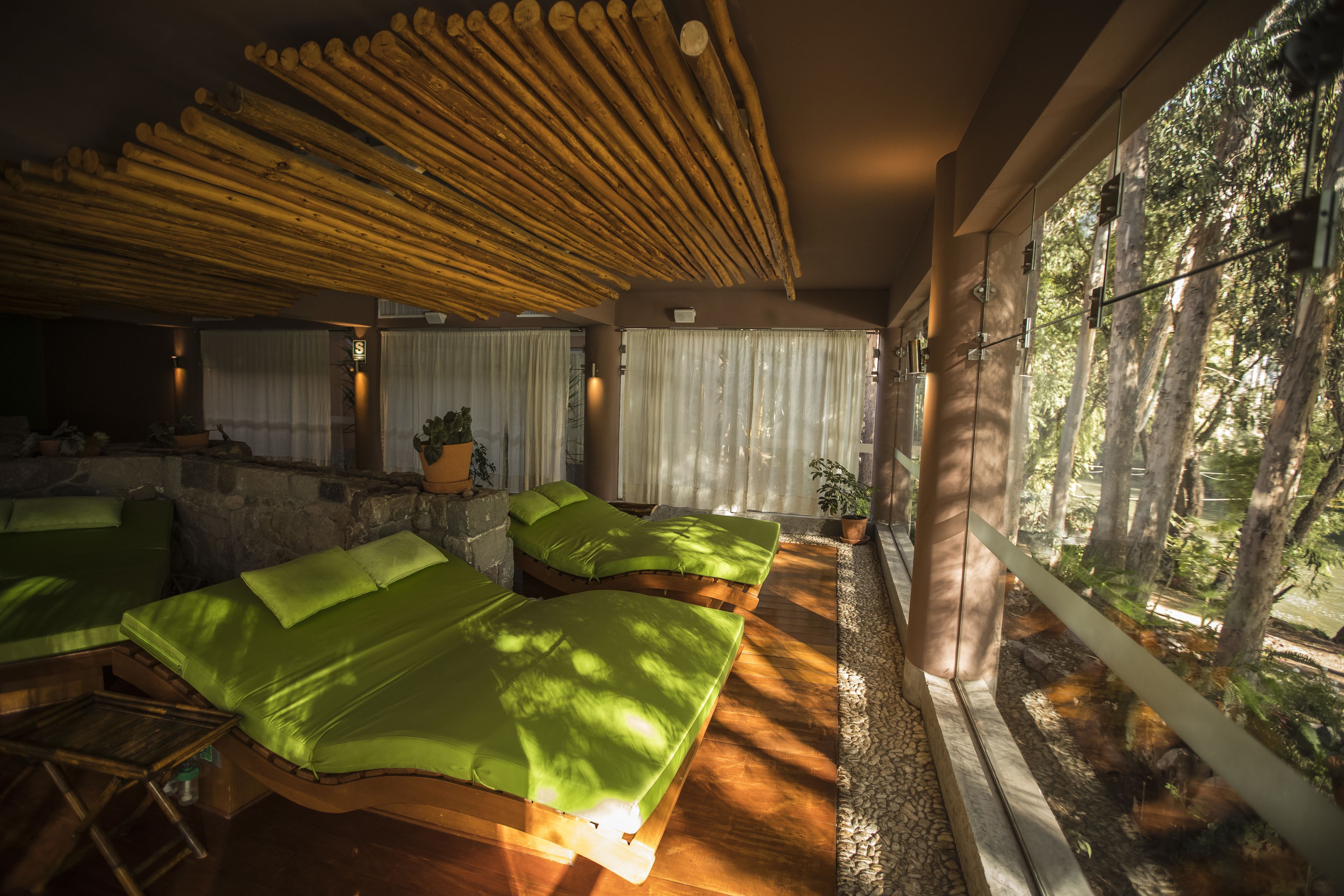 A spa room with wooden beds, a stone wall, and a view of trees through large windows.