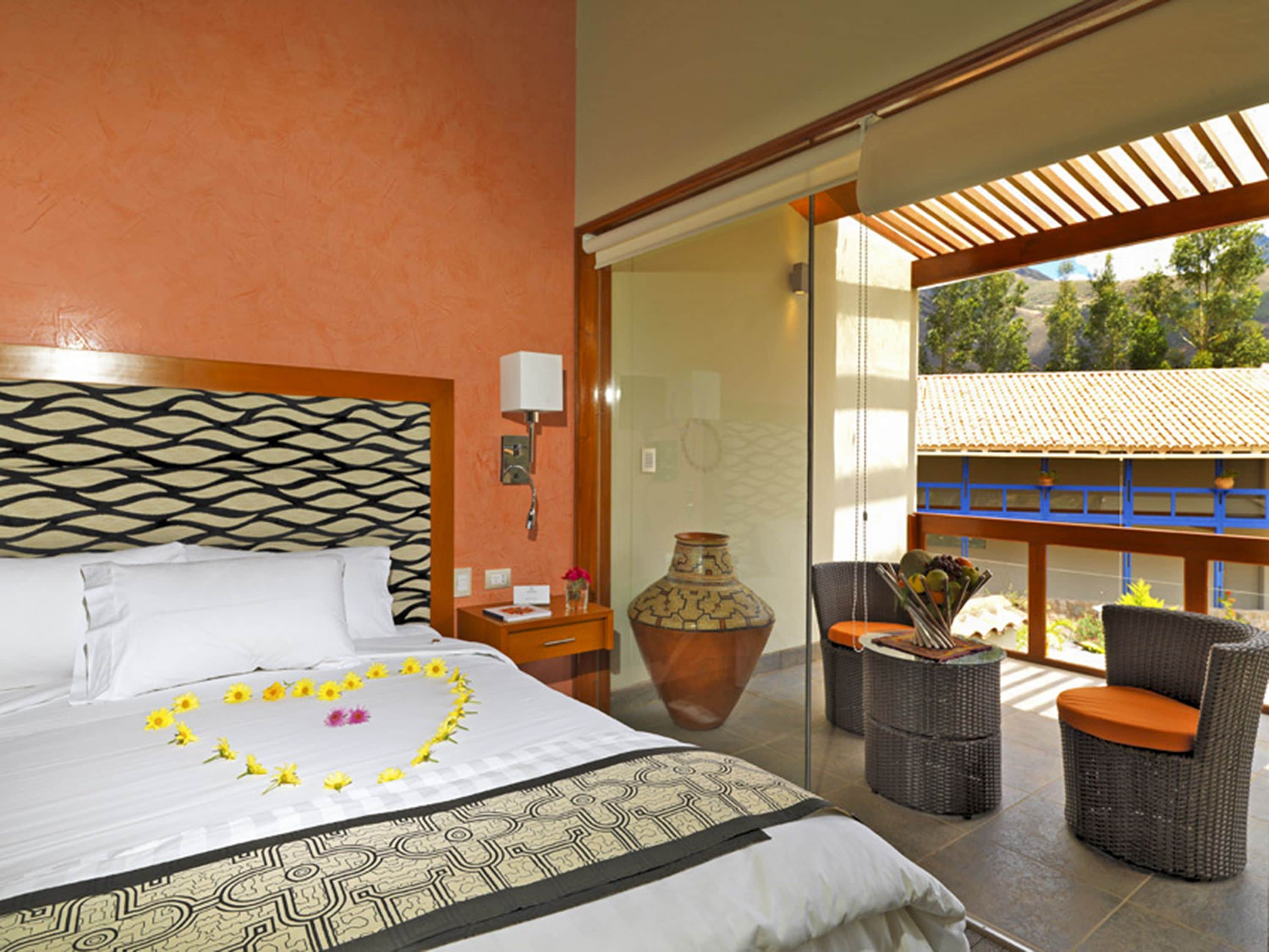 Habitación de hotel con cama, mesita de noche, florero, sillas de mimbre y vistas a zonas verdes.