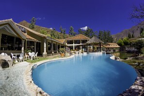 Una piscina con borde curvo, rodeada de mobiliario exterior y un edificio con grandes ventanales.