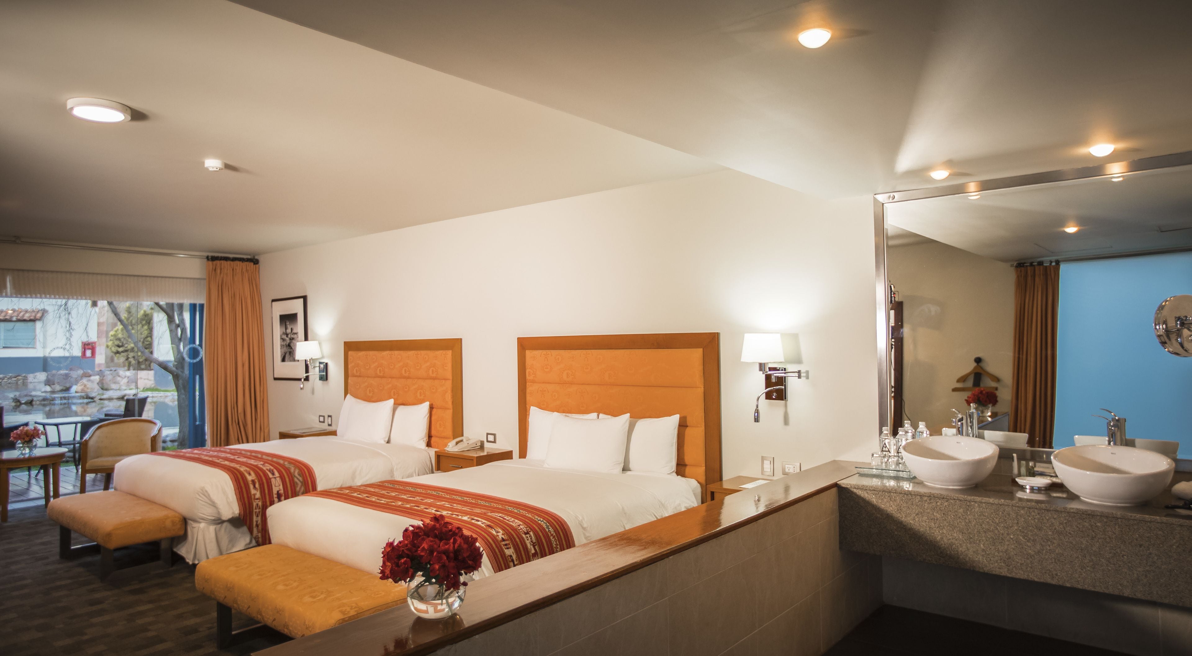 Habitación de hotel con dos camas, un baño con lavamanos y espejo, y una mesita con un jarrón de flores.