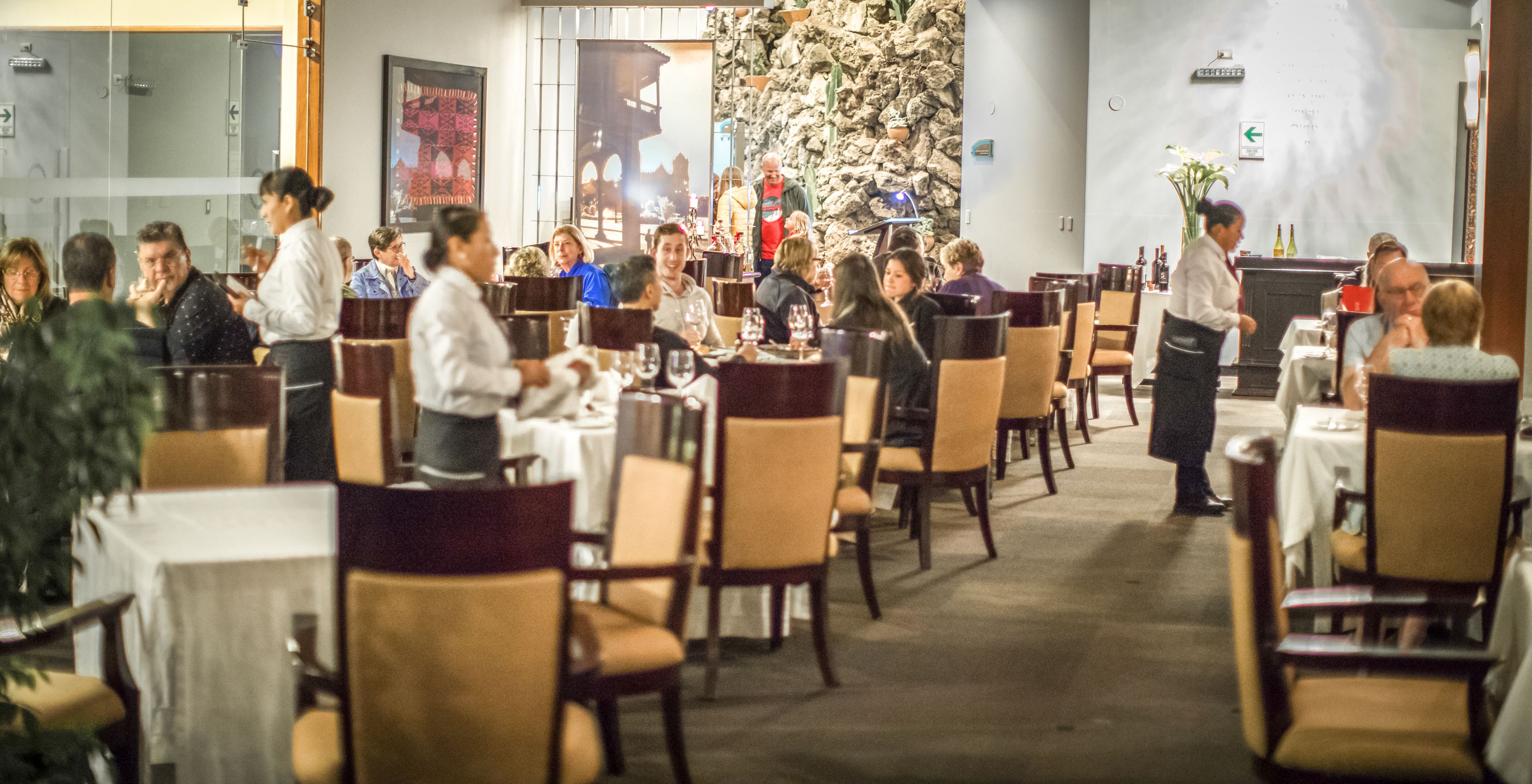 Un restaurante con varias mesas y sillas, un camarero atendiendo a los comensales y una pared decorativa.
