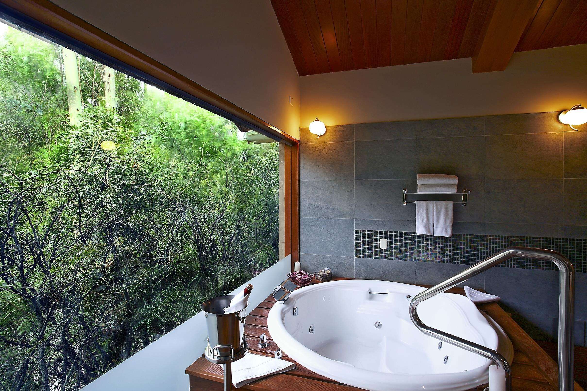 A modern bathroom with a large bathtub, a wooden ceiling, and a view of lush greenery outside.