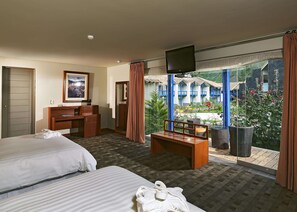 Habitación de hotel con dos camas, un escritorio, un televisor y un balcón con vistas a un edificio y áreas verdes.