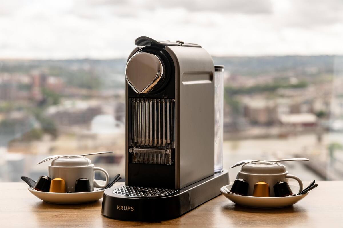 A coffee machine with two cups and a plate of coffee pods on a table with a city view in the background.