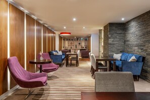 Un moderno hall de hotel con un panel de pared de madera, un detalle de pared de piedra, un área de estar central con sofás y sillones, y una estantería.