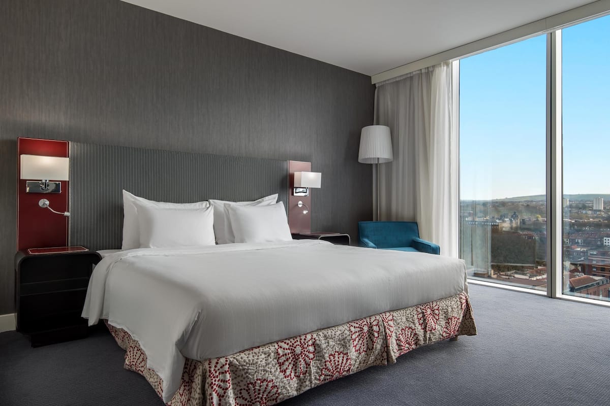A modern hotel room with a large bed, a nightstand, a lamp, and a view of the city through large windows.