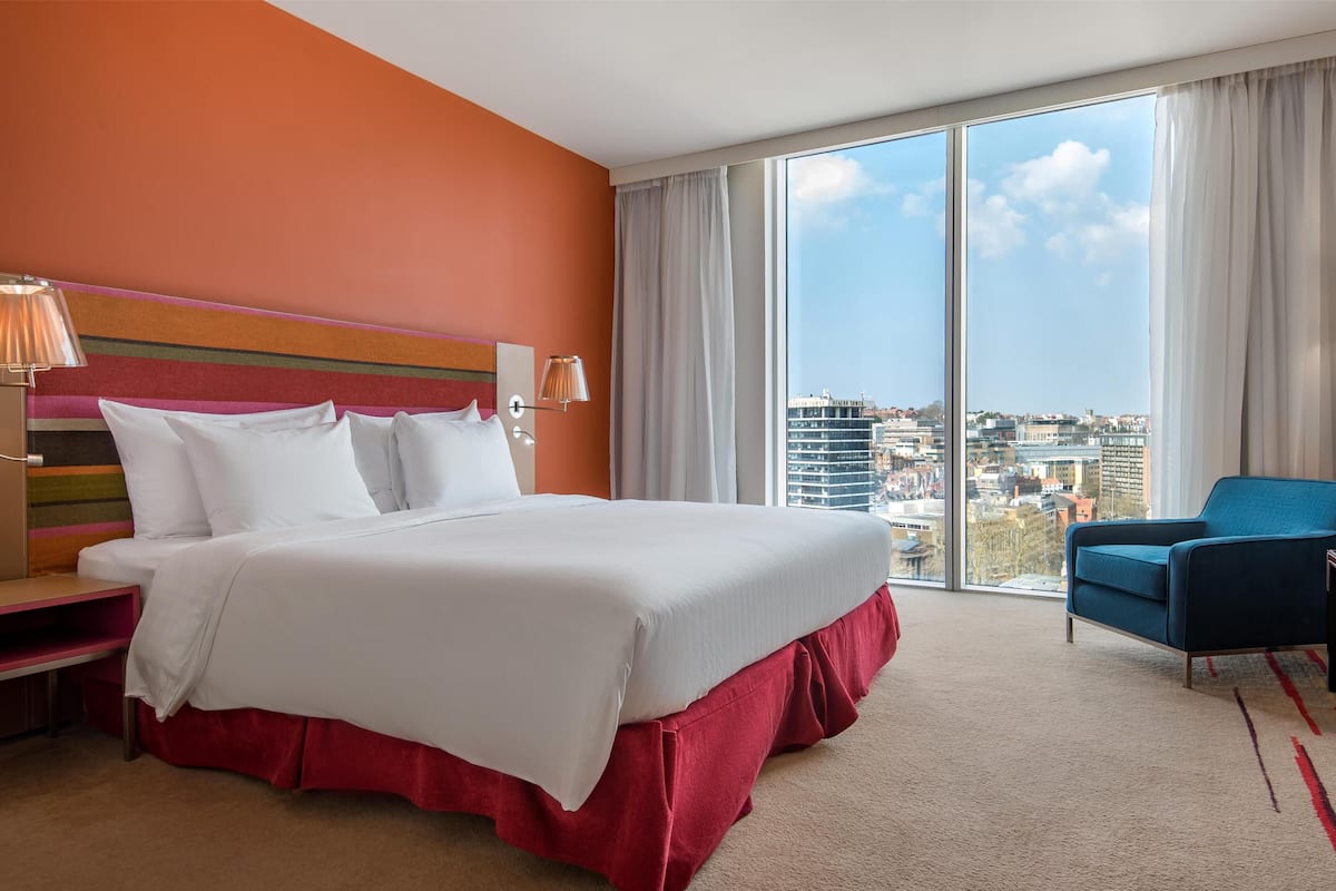 A hotel room with a large bed, a view of the city, and a blue armchair.