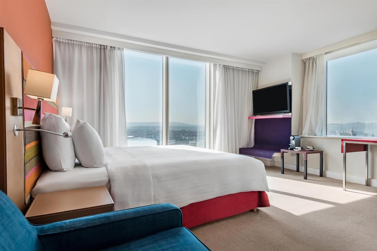 A hotel room with a large bed, a TV, a desk, and a view of the city.