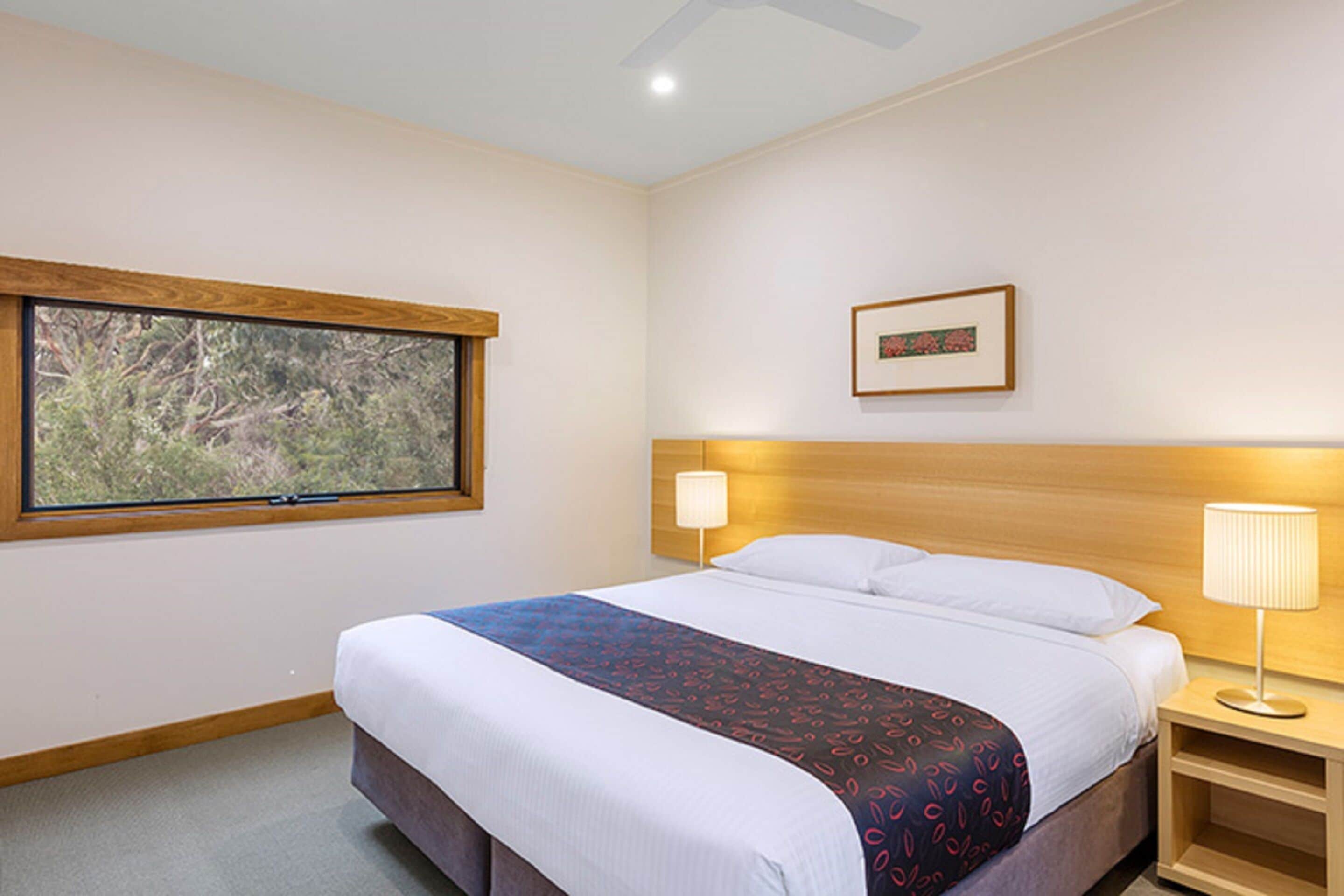 A hotel room with a large bed, a window with a view of trees, a bedside table with a lamp, and a framed picture on the wall.