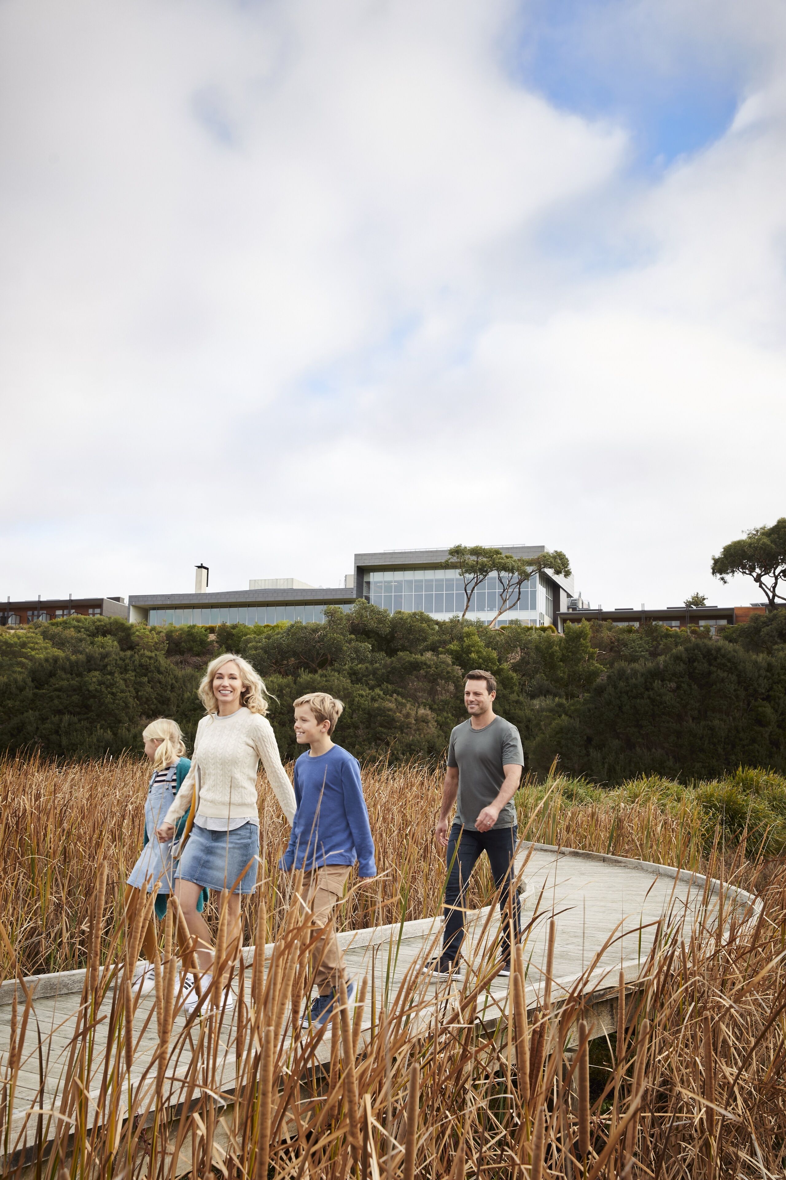 A family walking on a wooden boardwalk surrounded by tall grass and trees, with modern buildings in the background.