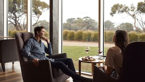 Un intérieur moderne avec une grande fenêtre donnant sur un espace verdoyant et des arbres. Deux personnes sont assises sur un canapé et une chaise, et une petite table se trouve entre elles, sur laquelle on peut voir un verre de vin et une assiette de nourriture.